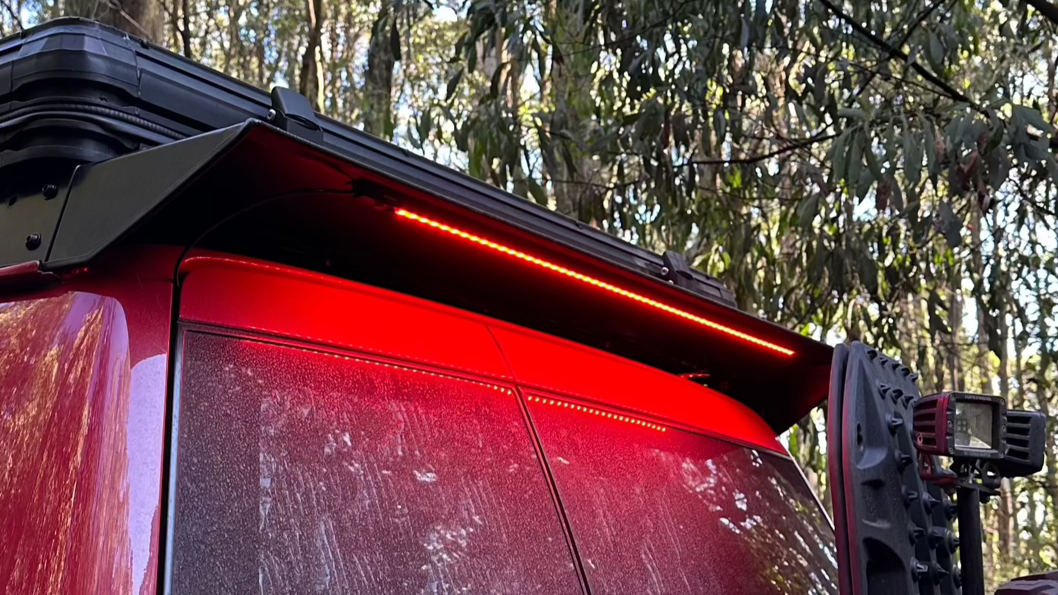This is an image of the rear side of a red 4WD featuring a black Tier One Metalworks roof rack with red LED strip lighting and off-road accessories in a forest setting