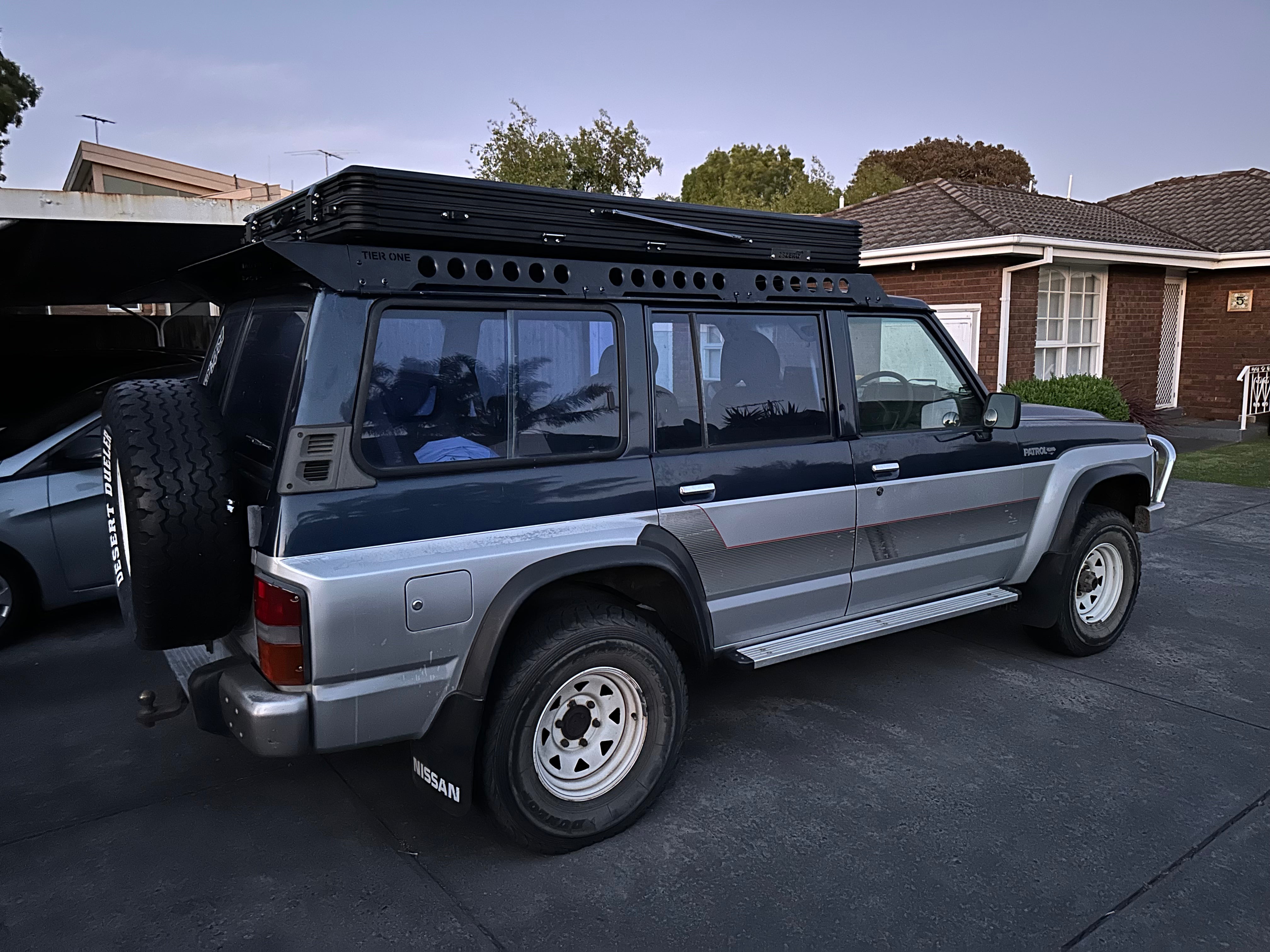 This is an image of a blue and silver Nissan Patrol with Tier One Metalworks roof rack and rear mounted spare Desert Dueler tyre
