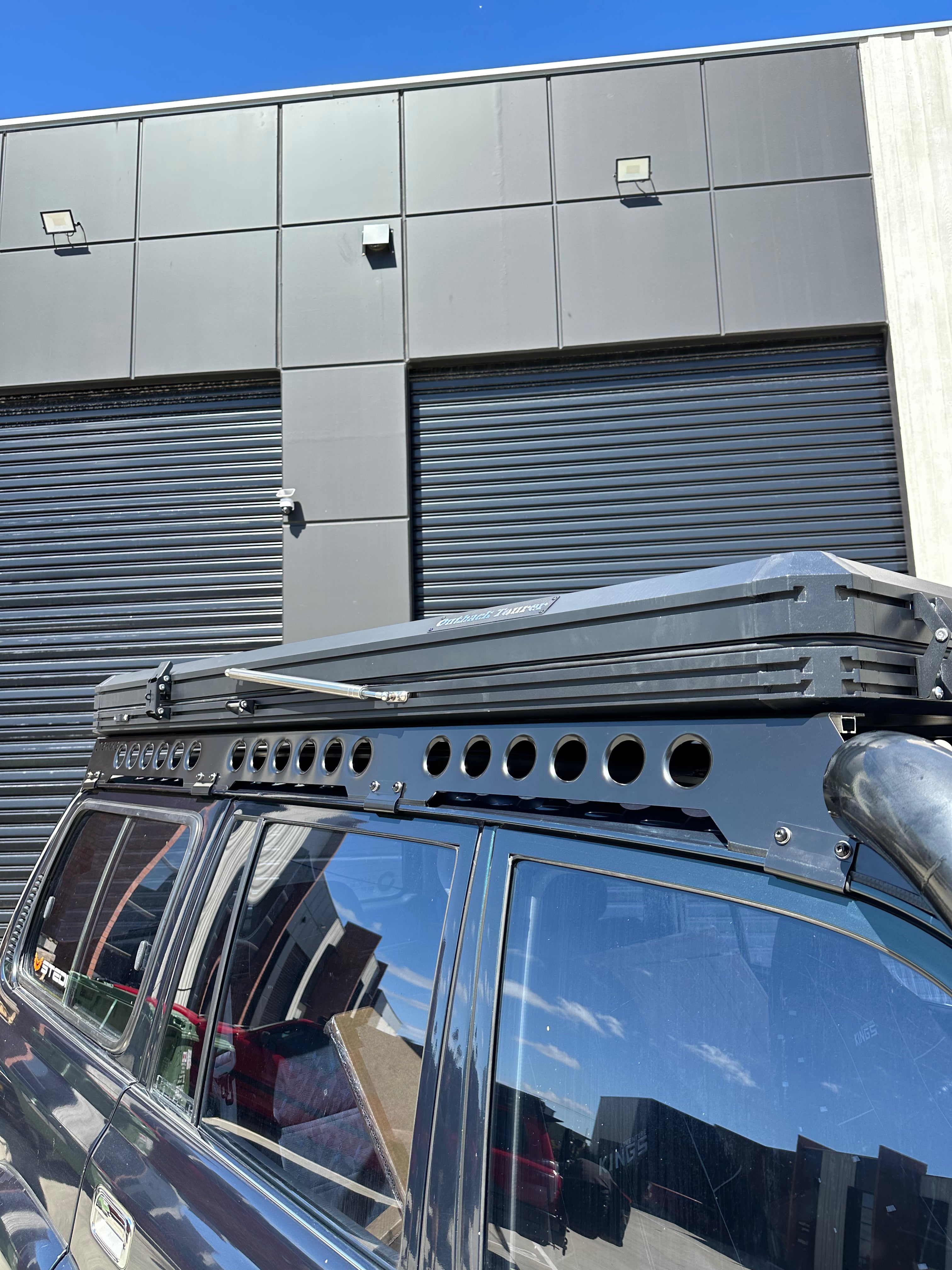 This is an image of a black 4WD with Tier One Metalworks black Roof Rack and Rails mounted, parked outside a warehouse