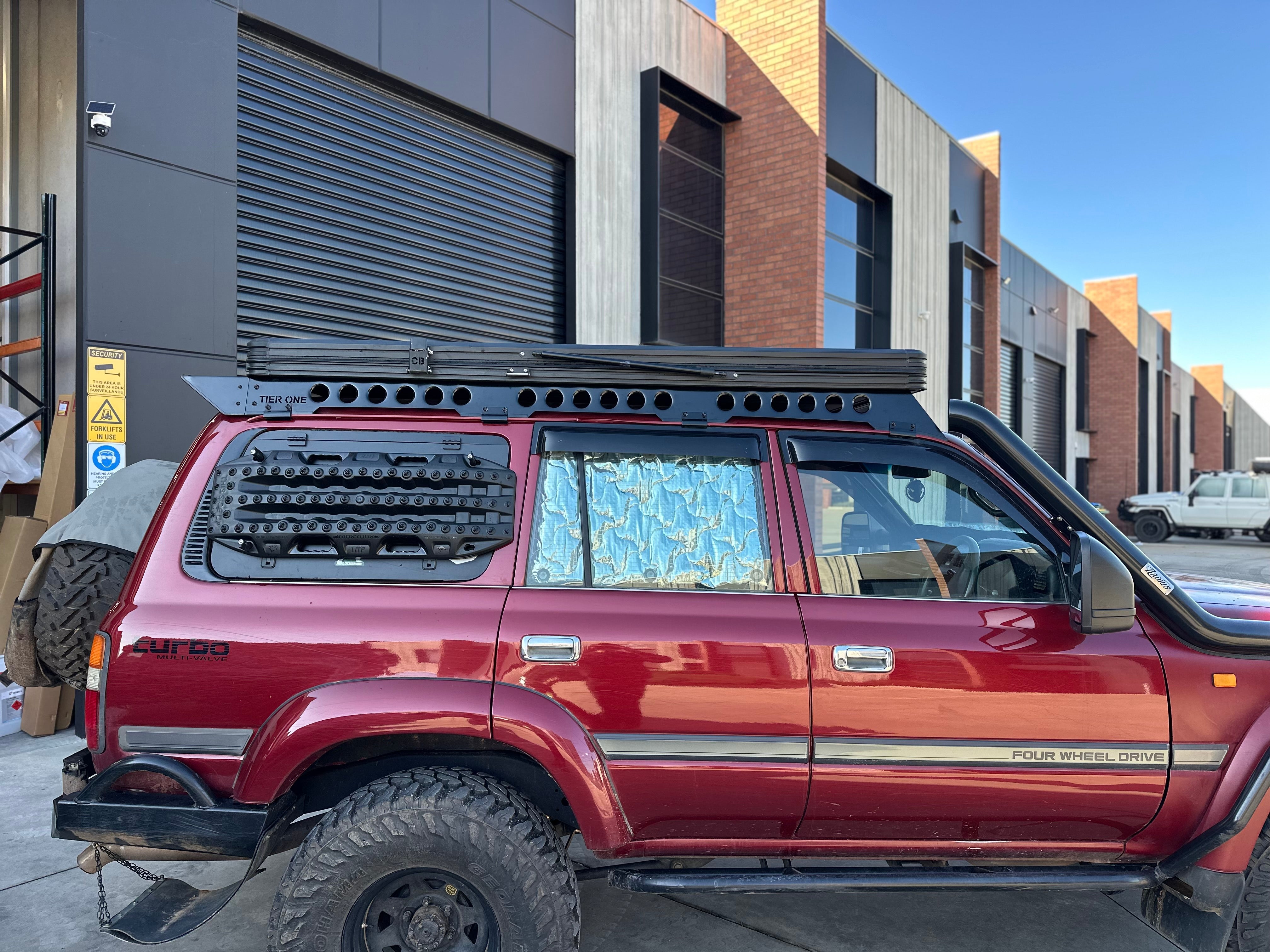 This is an image of a maroon 4WD Toyota Land Cruiser with Tier One Metalworks black roof rack and rails on an industrial background