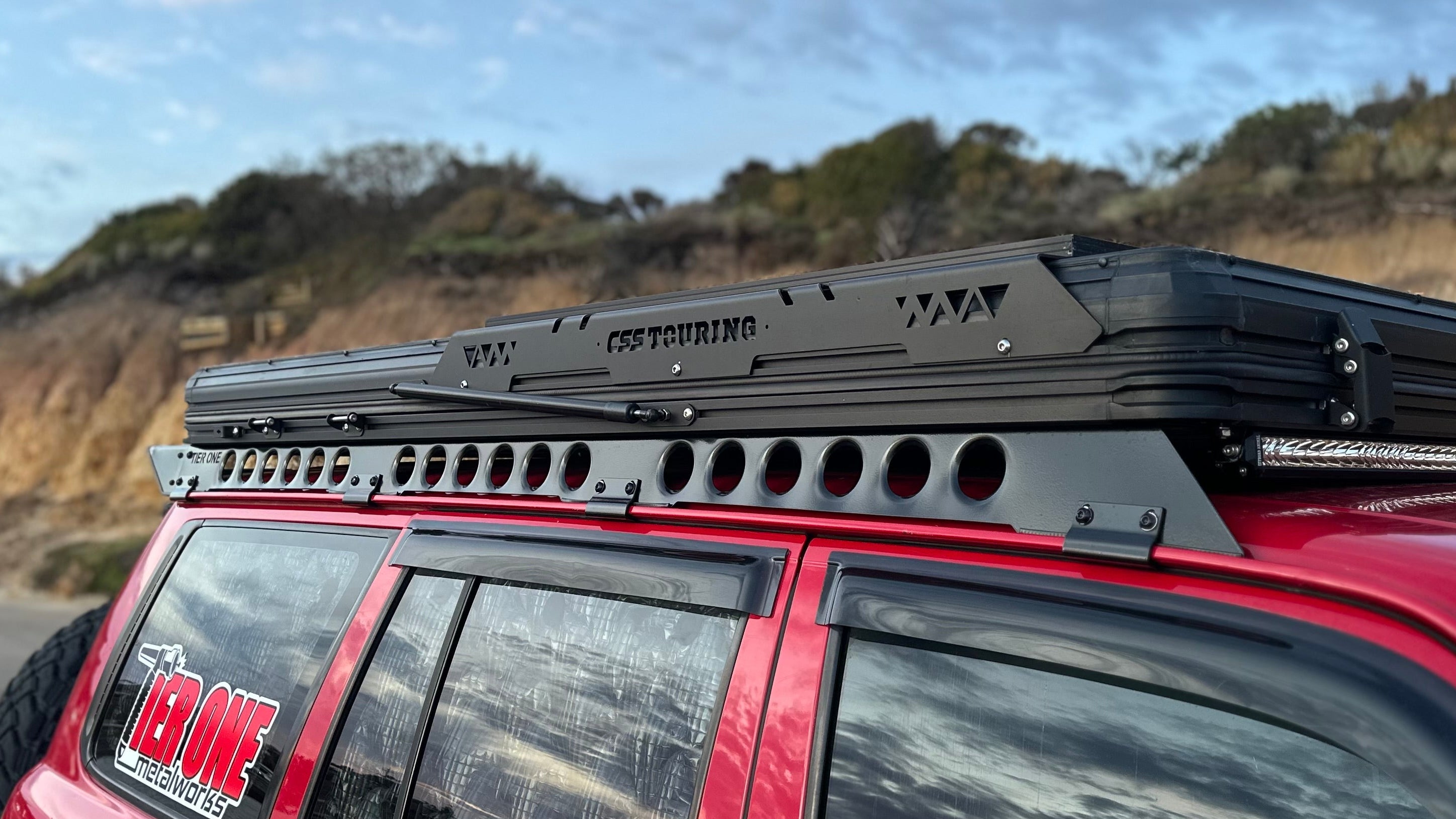 This is an image of a red 4x4 with a CSS Touring rooftop tent mounted on a Tier One Metalworks Roof Rack and Rails at sunset