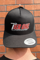 This is an image of a black snapback cap with red and white Tier One Metalworks logo embroidered on front, worn against a brick wall background