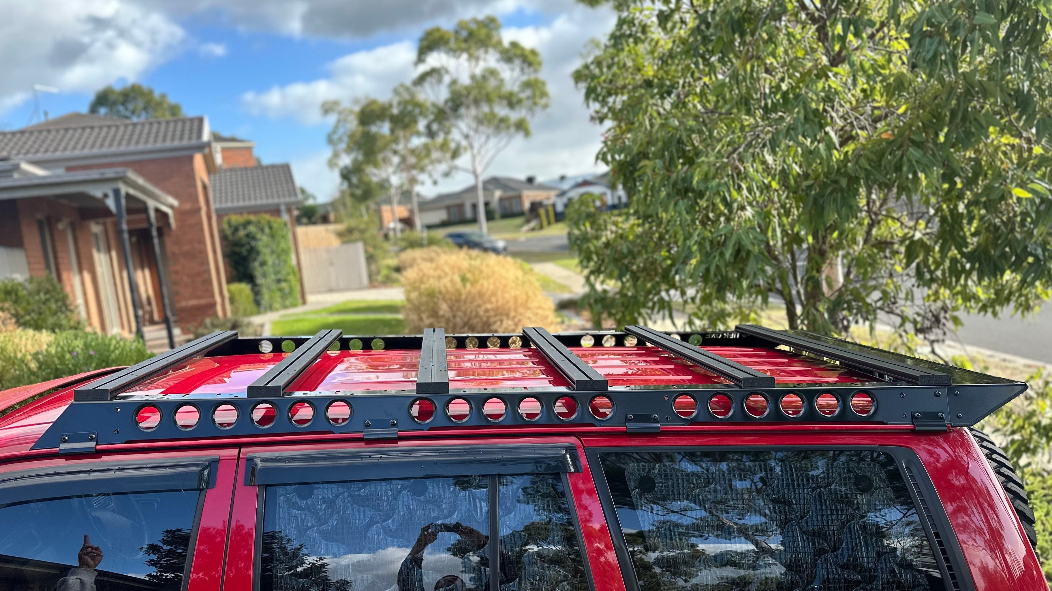 This is an image of a red SUV with a black Tier One Metalworks roof rack installed in a suburban street setting.