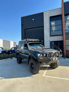 This is an image of a black Toyota Land Cruiser 4WD fitted with Tier One Metalworks roof racks and front bull bar parked outside a workshop
