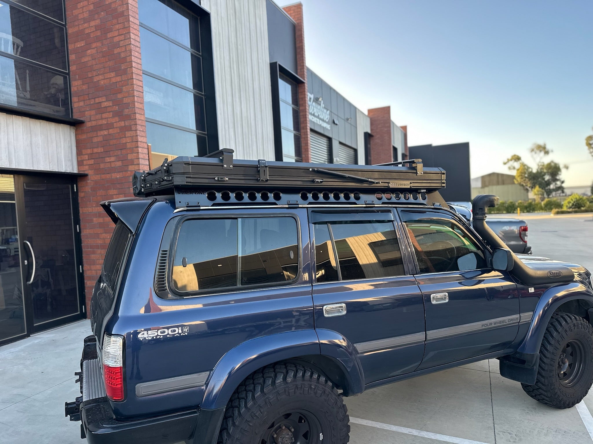 This is an image of a blue 4WD Toyota Land Cruiser 4500i with Tier One Metalworks roof racks and Safari snorkel in an industrial setting