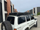 This is an image of a white Toyota Land Cruiser 80 Series with a black Tier One Metalworks roof rack and CSS Touring rooftop tent in an industrial area