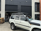 This is an image of a white 4WD SUV fitted with a Safari snorkel and a Tier One Metalworks roof rack with CSS Touring rooftop tent, parked outside an industrial garage.