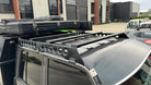 This is an image of a grey 4x4 vehicle featuring a Tier One Metalworks black roof rack and rails mounted on the roof in an industrial area