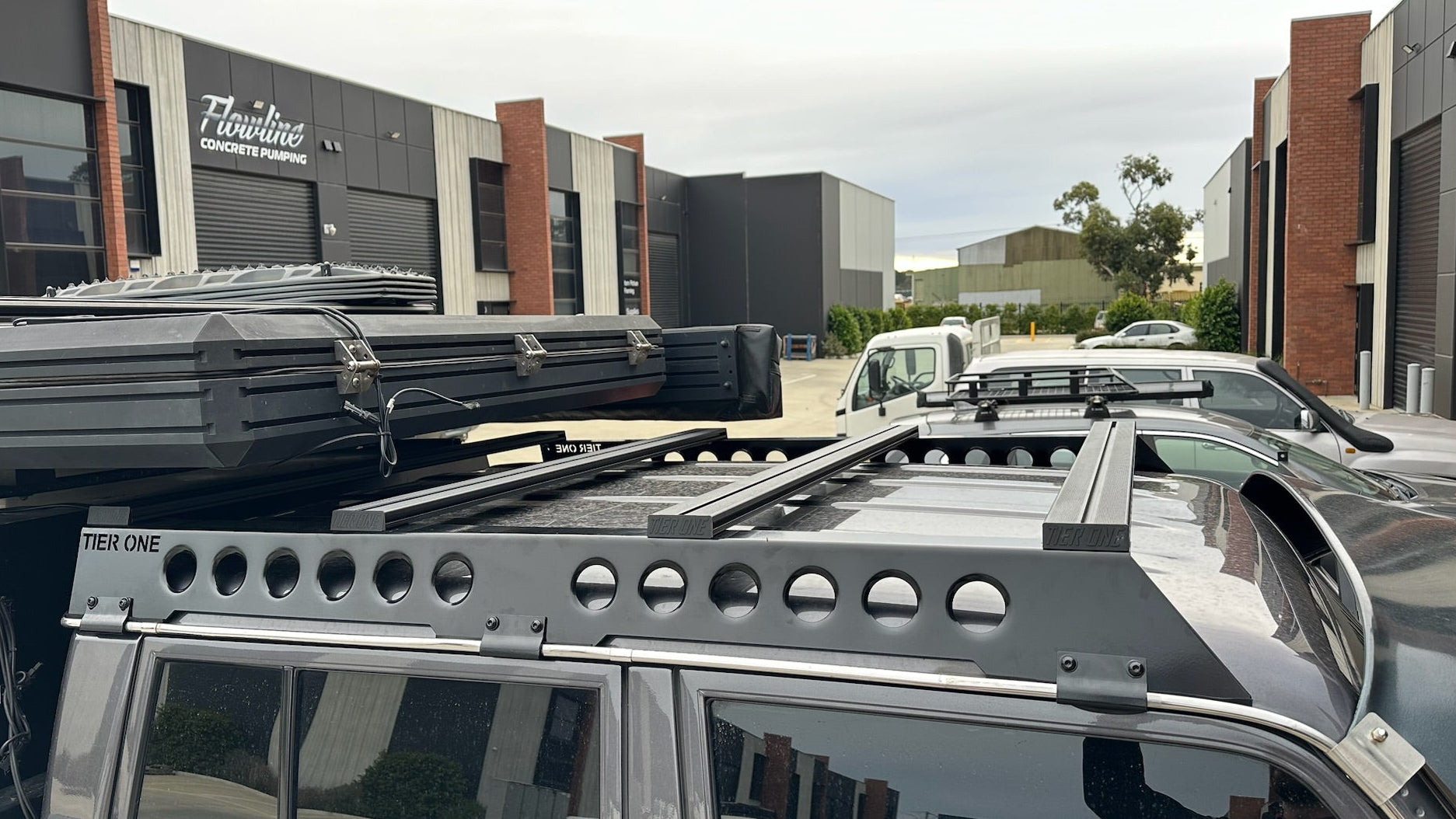 This is an image of a black Tier One Metalworks Roof Rack & Rails mounted on a dark 4WD with rooftop cargo and side windows in an industrial area.