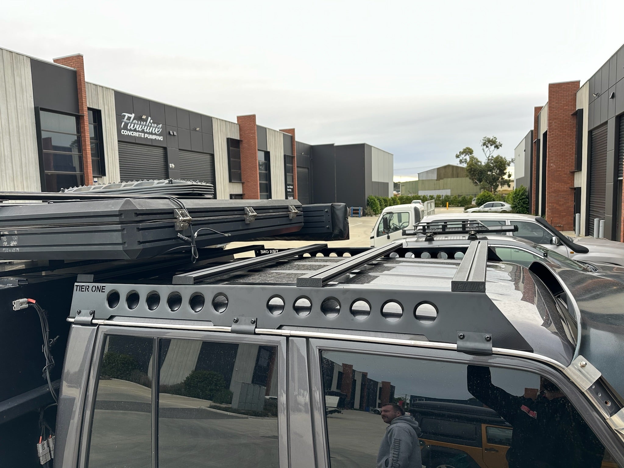 This is an image of a black Tier One Metalworks Roof Rack & Rails mounted on a dark 4WD with rooftop cargo and side windows in an industrial area.