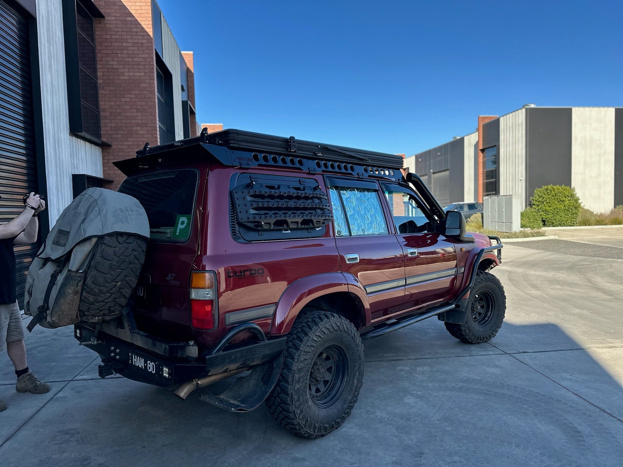 This is an image of a maroon Toyota Land Cruiser 80 Series with Tier One Metalworks black roof rack and side rails parked outside industrial buildings.