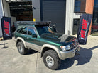 This is an image of a green and beige Nissan Patrol 4WD with Tier One Metalworks custom roof rack and snorkel parked outside a workshop.