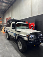 This is an image of a white Toyota Land Cruiser 70 Series with Tier One Metalworks roof racks and snorkel in a workshop setting.
