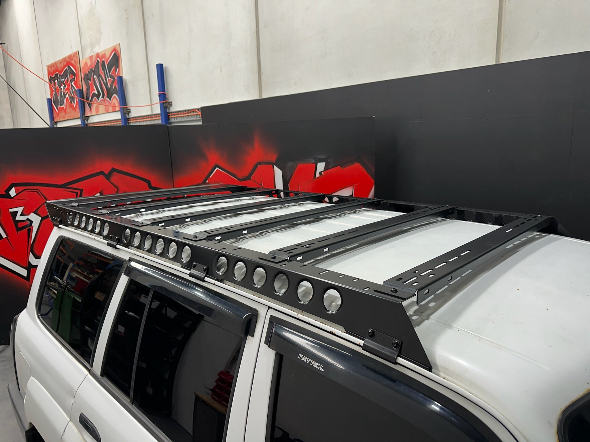 This is an image of a black Tier One Metalworks roof rack mounted on a white Nissan Patrol 4x4 in a workshop