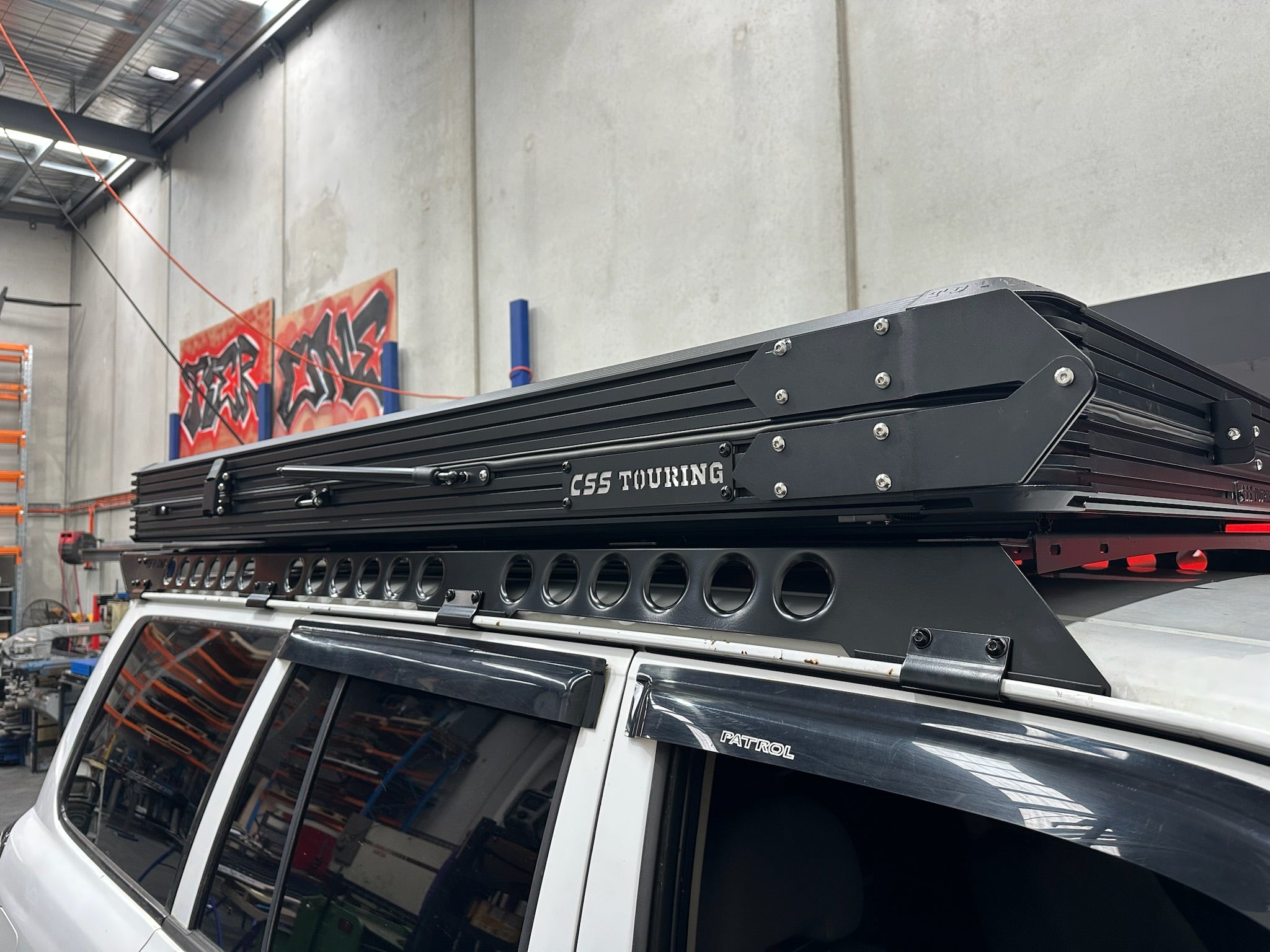 This is an image of a white Nissan Patrol with a CSS Touring rooftop tent mounted on a Tier One Metalworks black roof rack inside a workshop