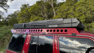 This is an image of a red 4WD with a Tier One Metalworks roof rack and a black CSS Touring rooftop tent on top in an outdoor setting