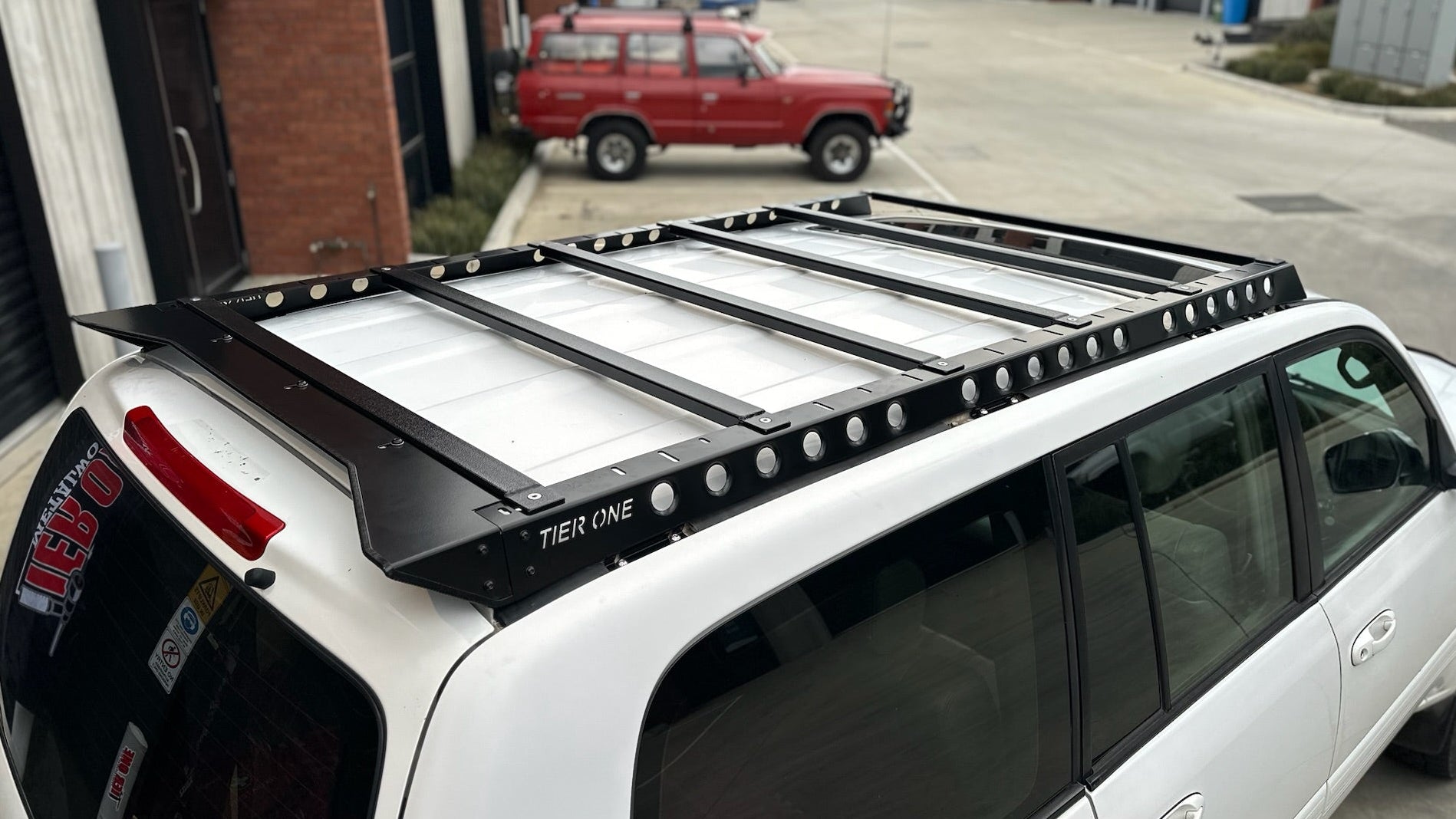 This is an image of a white Toyota 4x4 fitted with a black Tier One Metalworks roof rack in an industrial parking lot