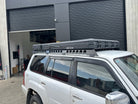 This is an image of a white Nissan Patrol with black Tier One Metalworks Roof Racks & CSS Touring rooftop tent in a workshop