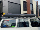 This is an image of a white 4WD with a Tier One Metalworks black roof rack and rooftop tent parked outside a workshop