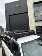 This is an image of a white 4x4 vehicle with Tier One Metalworks black roof racks and a closed roof top tent mounted on top, parked in an industrial area.