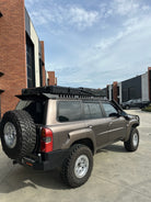 This is an image of a brown Nissan Patrol fitted with Tier One Metalworks Roof Racks & Rails and rugged off-road tyres at an industrial site.