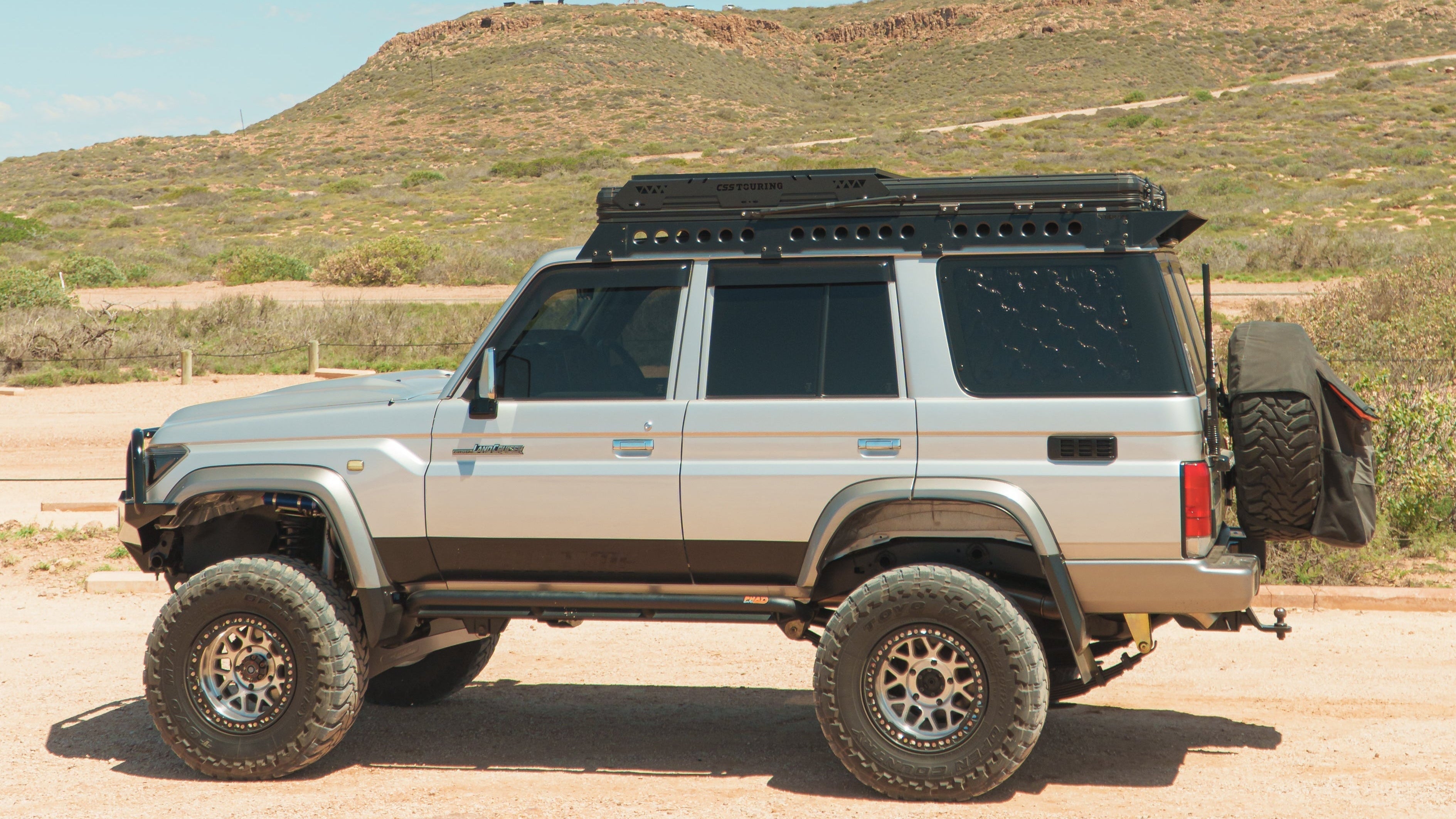 This is an image of a silver Toyota Land Cruiser with Tier One Metalworks Roof Racks & Rails and a CSS Touring rooftop tent on a dirt road