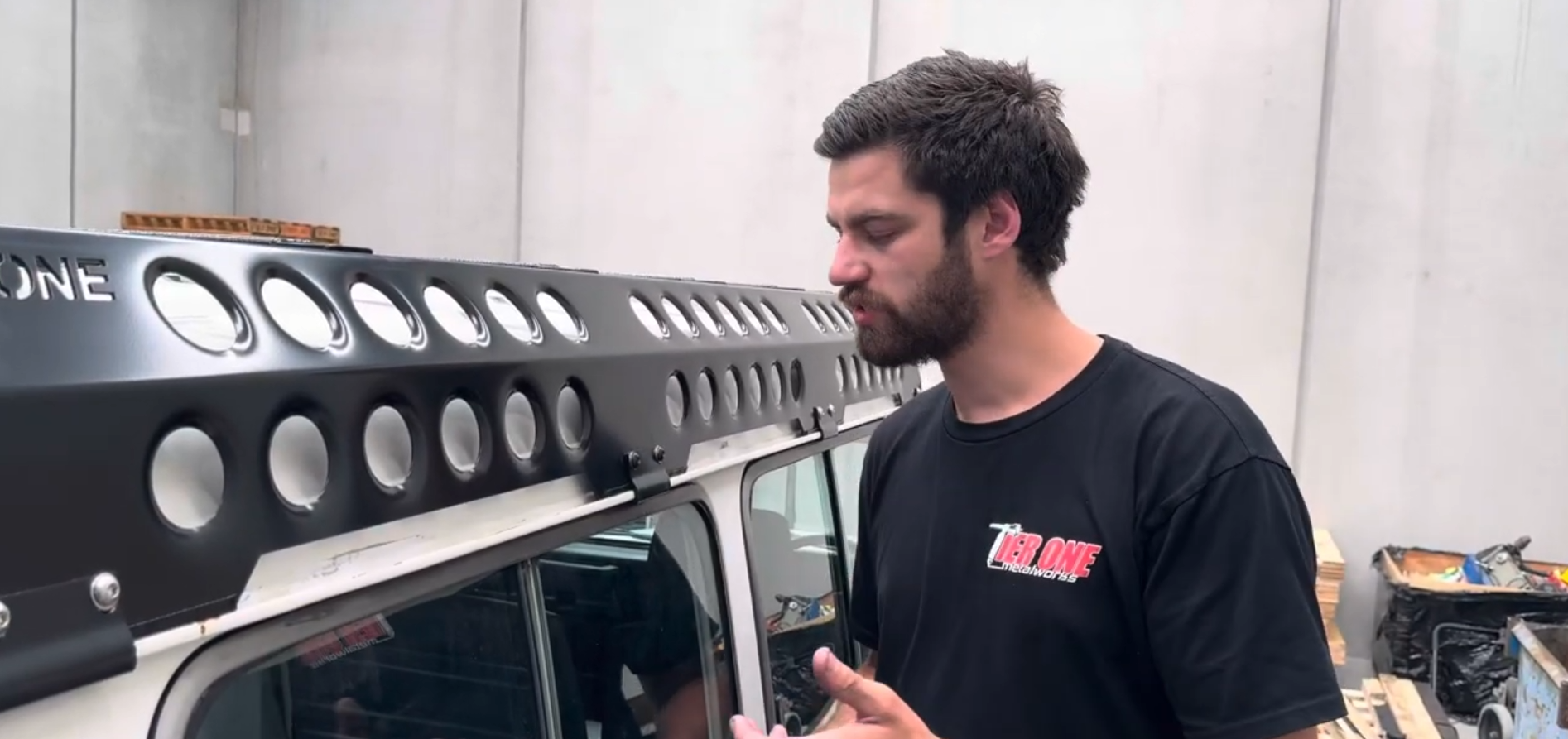 This is an image of a man inspecting a black Tier One Metalworks roof rack mounted on a white vehicle inside a workshop.