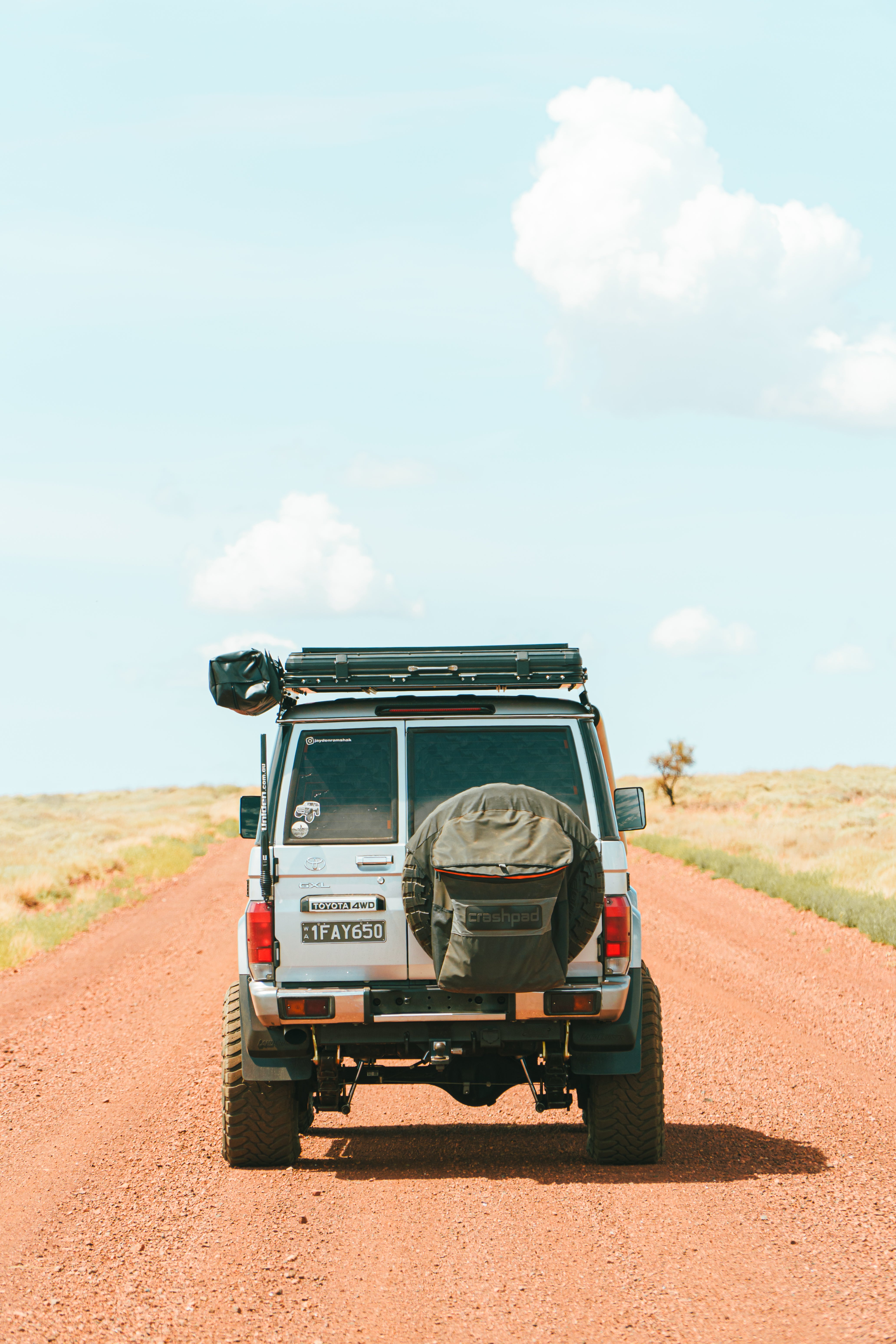 This is an image of a white Toyota Landcruiser 76 Series driving on a red dirt road in outback Australia featuring a black Tier One Metalworks roof rack with rear wing and a spare tyre cover