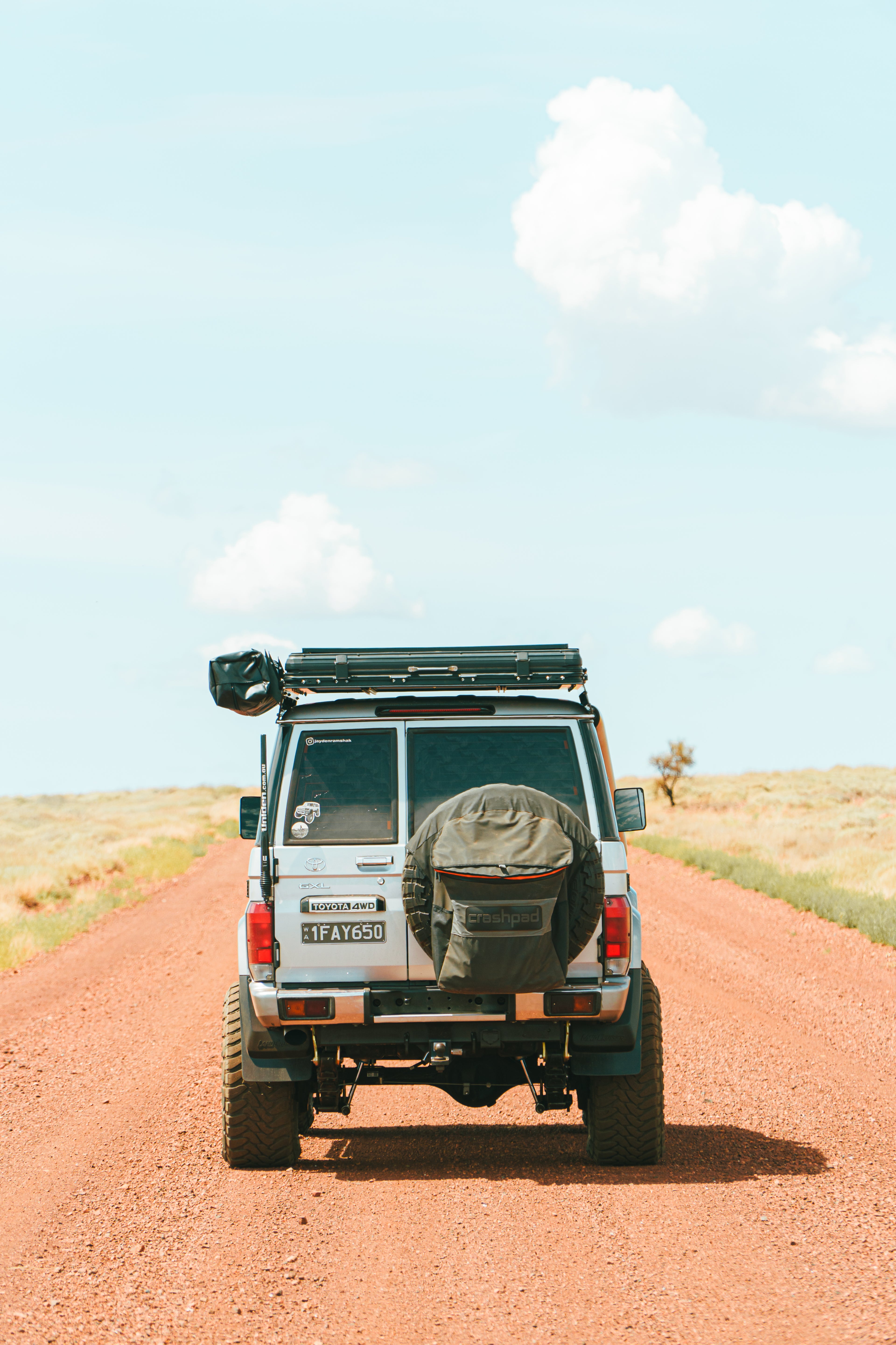 This is an image of a white Toyota Landcruiser 76 Series driving on a red dirt road in outback Australia featuring a black Tier One Metalworks roof rack with rear wing and a spare tyre cover